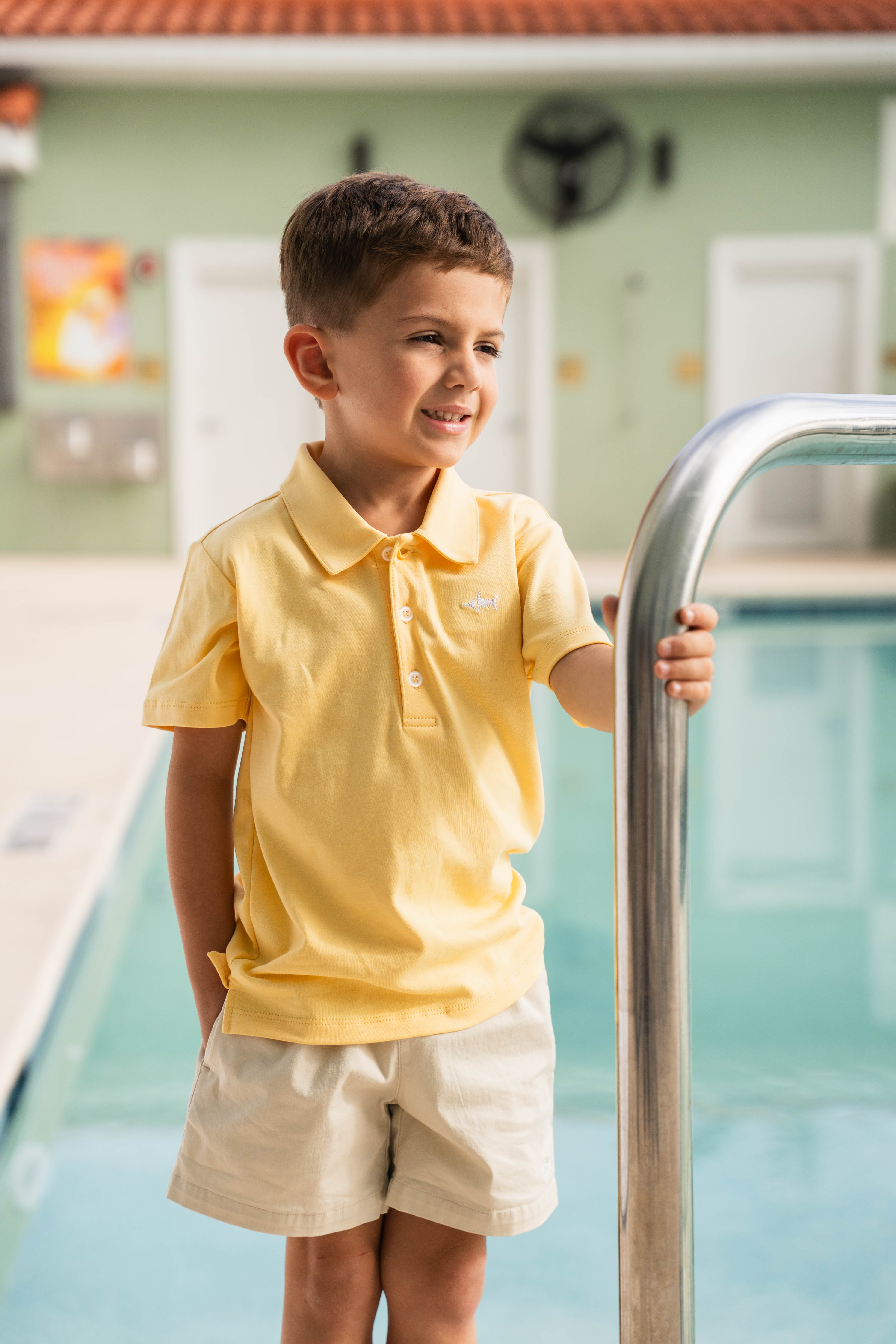 Young boy in a yellow polo shirt and beige shorts standing by a pool.