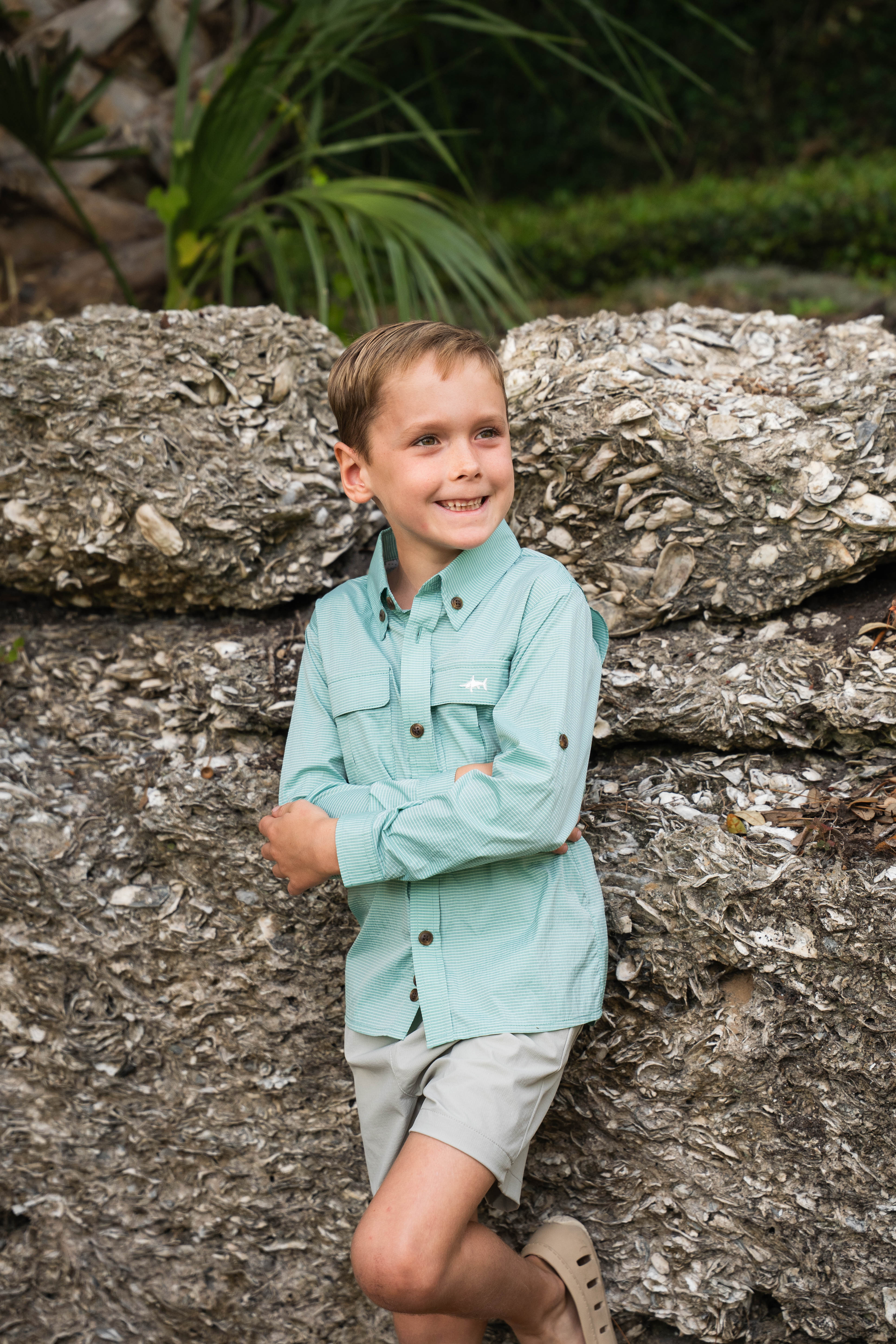 Young boy in a light blue shirt standing among rocks with greenery in the background