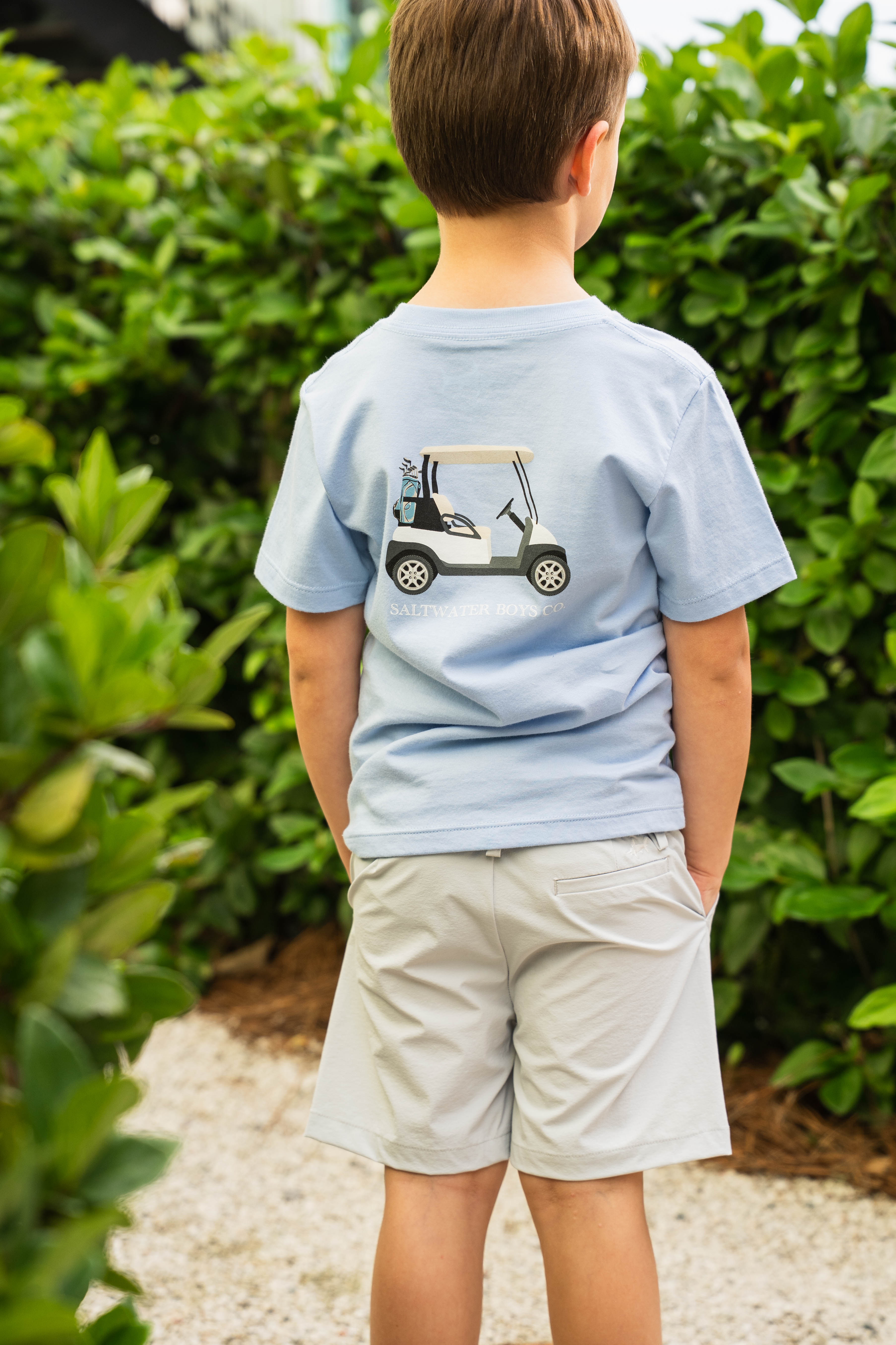 Child wearing a light blue t-shirt with a golf cart graphic, standing in front of green foliage.
