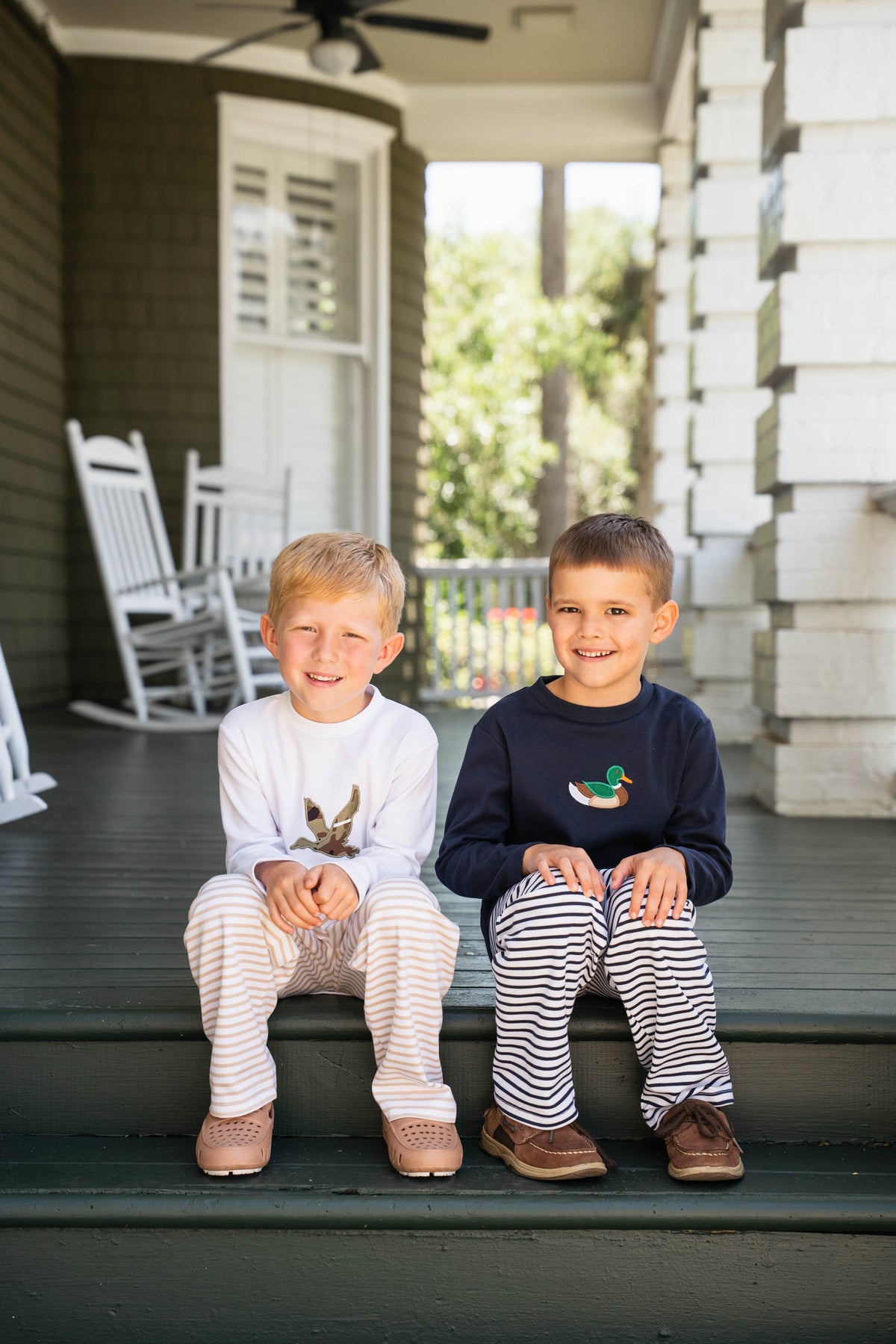 Two young boys sitting side-by-side on the porch steps of a home, wearing matching long-sleeve pajamas. The boy on the left wears a white pajama top with a brown bunny graphic and light beige striped pajama pants.