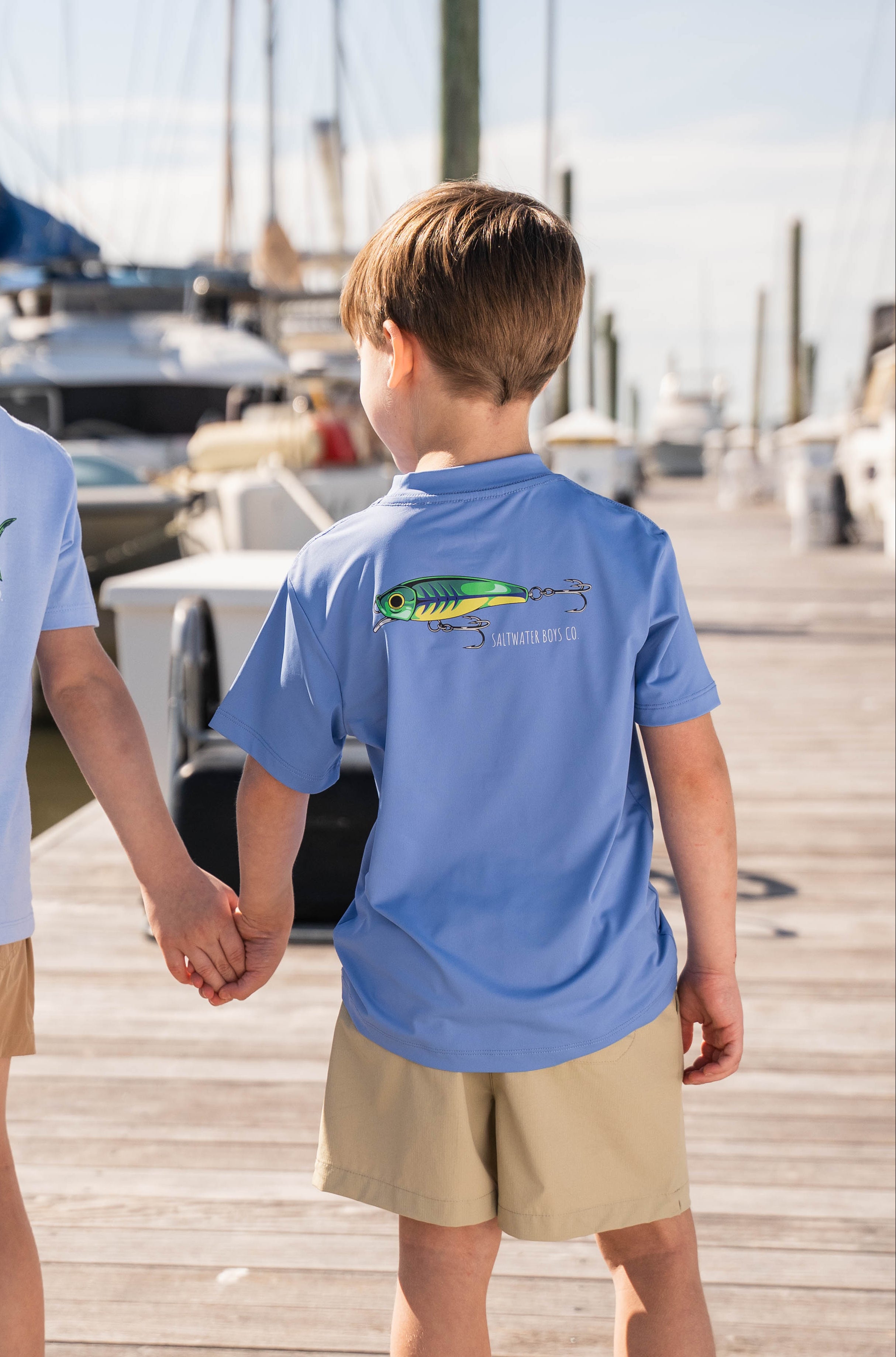 Two children holding hands on a dock wearing matching blue t-shirts with a fish graphic.
